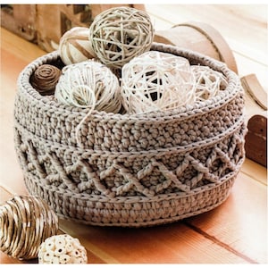 Op de afbeelding: Een beige gehaakte mand met een geweven patroon, gevuld met witte en bruine decoratieve ballen en garen.