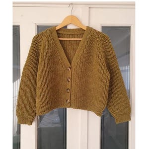 May include: Olive green, chunky knit cardigan with a V-neck and three brown buttons. The sweater is displayed on a wooden hanger. The sleeves are long and the knit has a ribbed texture. A cozy, handmade garment.