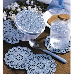 May include: Four blue crocheted doilies, one on a white and blue box, one under a glass of water with ice, and two on a wooden table.