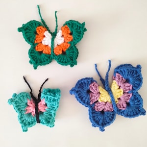 May include: Three crocheted butterflies in different colors and patterns. The butterflies are green with orange and white accents, blue with pink and yellow accents, and teal with pink accents.