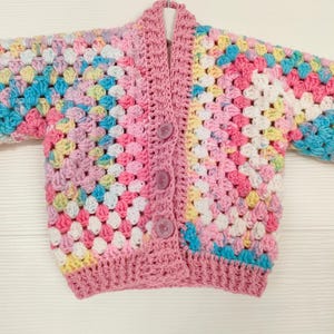 以下が含まれることがあります： カラフルなグラニー・スクエア模様のピンクのクロシェ編みカーディガン。カーディガンはボタン留めと長袖です。