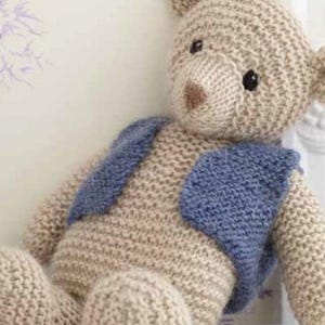 Puede incluir: Un oso de peluche tejido con un chaleco tejido azul. El oso est&aacute; sentado sobre una superficie blanca.