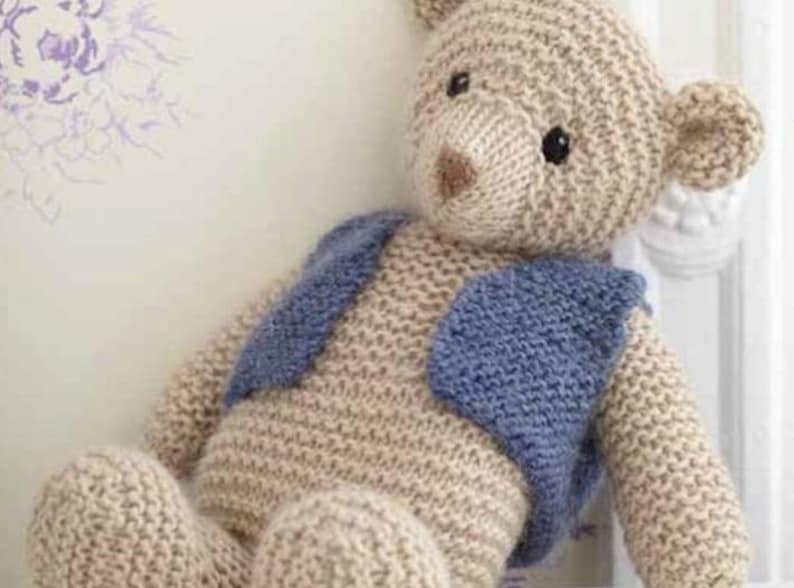 Puede incluir: Un oso de peluche tejido con un chaleco tejido azul. El oso est&aacute; sentado sobre una superficie blanca.