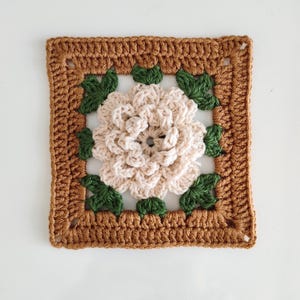 May include: A crocheted square with a white flower in the center surrounded by green leaves. The square has a brown border.