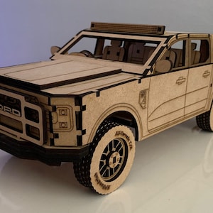 Puede incluir: Maqueta de madera detallada de una camioneta Ford. El modelo presenta intrincados detalles cortados con láser, incluido el logotipo de Ford en la parte delantera, neumáticos todoterreno y una barra de luces en el techo. El camión está hecho de madera clara.