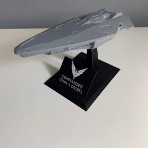 Può includere: Un modello di nave spaziale stampato in 3D di colore grigio su un supporto nero. Il supporto ha un logo bianco con il testo "Commander Chin x Diesel".