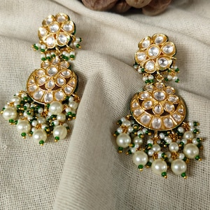 Può includere: Orecchini color oro con un design floreale e perle pendenti. Gli orecchini presentano pietre rotonde e trasparenti e accenti verdi. La parte inferiore degli orecchini ha perle bianche e perline verdi. Gli orecchini sono esposti su un tessuto color crema.