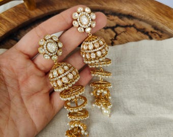 May include: Gold-toned chandelier earrings with a floral design at the top and a dangling, layered bell shape. The earrings are adorned with clear gemstones and small, white pearls, creating a luxurious look.