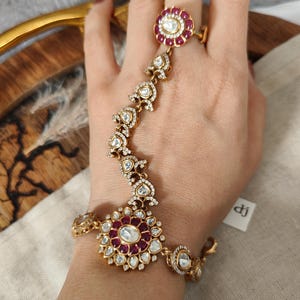 May include: Gold-toned hand chain bracelet and ring set. The jewelry features a floral design with red and clear gemstones. The ring and bracelet are connected by a delicate chain adorned with small, sparkling accents. The bracelet wraps around the wrist.