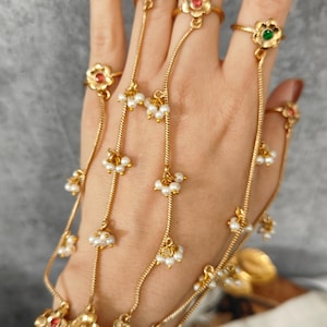 May include: Gold-toned hand jewelry featuring flower-shaped rings connected by delicate chains. The rings are adorned with red and green gemstones and small white pearls. The jewelry is designed to be worn across the hand.
