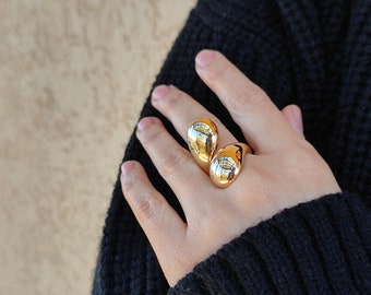 Könnte beinhalten: Ein goldener Ring mit zwei großen, runden, glatten, verbundenen Kugeln. Der Ring befindet sich an einem Finger.