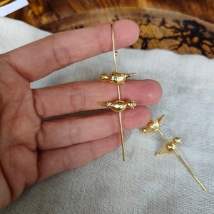 May include: Gold-toned earrings with a dangling design featuring three small bird charms. The birds are facing the same direction and have a textured surface.