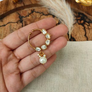 May include: A gold-toned nose ring featuring a circular band adorned with five clear, irregularly shaped stones along the upper curve and a single white pearl drop pendant accented by three tiny white beads at the base of the jewellery piece.