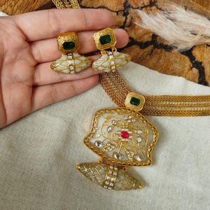 May include: A gold-toned necklace and earring set featuring multi-strand chains. The pendant and earrings showcase white, lattice-patterned stones with gold accents, rectangular green stones, and a central red stone on the main pendant piece.