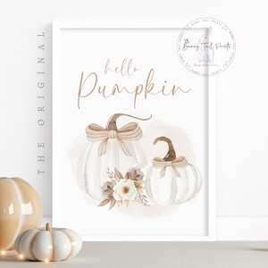 May include: A printable art print with the text "hello pumpkin" in brown script font. Two white pumpkins with brown stems and bows are illustrated on a pink watercolour background.