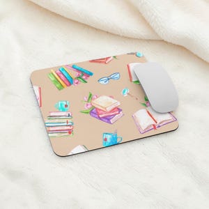 May include: A beige rectangular mousepad featuring a watercolor design of books, teacups, eyeglasses, and flowers. A white computer mouse rests on the right side of the pad. The design includes stacks of books in various colors.