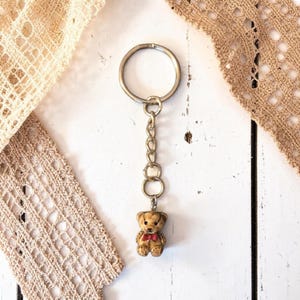 May include: A keyring with a small teddy bear charm wearing a red bow tie. The bear is attached to a chain and a silver-coloured keyring. The background features white wooden planks and beige lace.