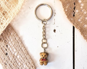 May include: A keychain featuring a small, detailed teddy bear charm with a red bow tie. The bear is attached to a chain and a silver-colored keyring. The background includes white wooden planks and beige lace.