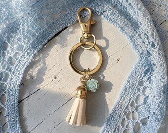 May include: A gold-colored keychain with a clasp, a ring, a cream-colored tassel, and a light blue rose charm. The keychain is set against a light blue lace fabric and a white wooden surface.