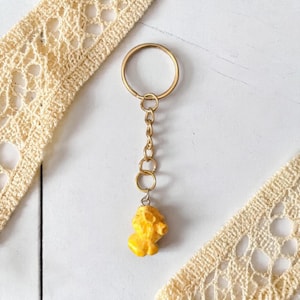 Op de afbeelding: Een goudkleurige sleutelhanger met een kleine, gele, popcornvormige bedel. De sleutelhanger heeft een cirkelvormige ring, een ketting en een popcornbedel. De achtergrond is wit met crèmekleurig kant.