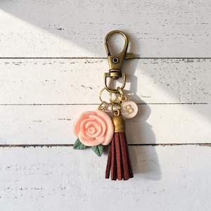 Könnte beinhalten: Ein Schlüsselanhänger mit einem bronzefarbenen Karabiner und Ringen. Er hat eine rosa Rose, eine braune Quaste und einen weißen Anhänger mit dem Buchstaben "B". Der Schlüsselanhänger befindet sich vor einem weißen Holzgrund.