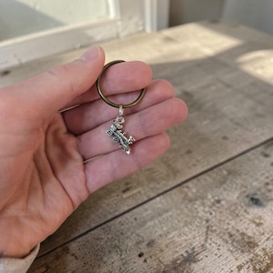 May include: A circular bronze-toned metal keyring featuring a small, detailed silver-toned metal charm shaped like a vintage steam locomotive train. The charm is attached to the ring by a small jump ring, showcasing intricate wheels and engine details.