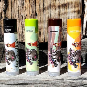 May include: Six different flavored lip balms with a safety seal. Each balm features a cartoon illustration of a chicken and a creature with a flower crown. The flavors are Hot Moss Heifer, Tear Seal, and four others.