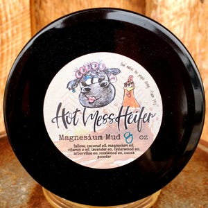 May include: A black jar with a white label that says "Hot Mess Heifer Magnesium Mud 8 oz". The label also includes a cartoon cow and chicken with the text "God makes his people strong - Psalms 141".