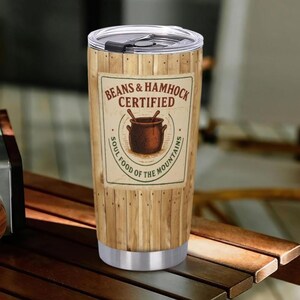 May include: A stainless steel travel tumbler with a wooden plank design. The tumbler features a label that reads "Beans & Hammock Certified" with a graphic of a pot and the text "Soul Food of the Mountains."