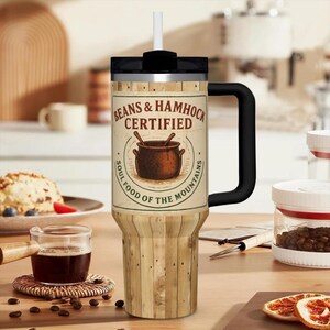 May include: A large, insulated tumbler with a black handle and lid, and a white straw. The tumbler features a wooden panel design and a label that reads "BEANS & HAMMOCK CERTIFIED" with a graphic of a pot. The text "SOUL FOOD OF THE MOUNTAINS" is also included.