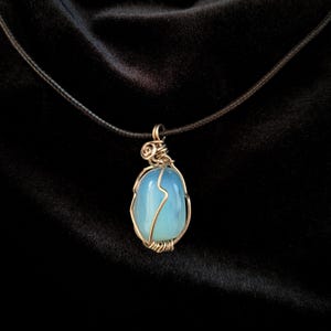 May include: A wire-wrapped pendant necklace featuring a light blue, oval-shaped stone. The stone is encased in a gold-colored wire, suspended from a thin, black cord. The background is a dark, textured fabric.