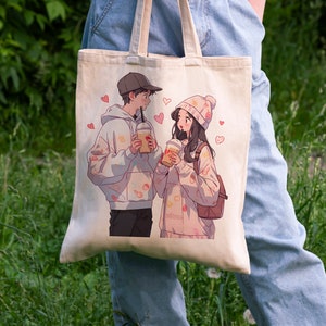 May include: A white canvas tote bag with a printed design of two cartoon characters holding beverages. The characters are a man with a black hoodie and a woman with a pink hoodie. The design includes pink hearts and other small details.