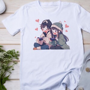 May include: A white t-shirt with a graphic print of two anime characters, a boy and a girl, playing video games. The boy is wearing a blue and white hat and a brown sweater. The girl is wearing a green sweater and a white hat. The graphic is surrounded by pink hearts.