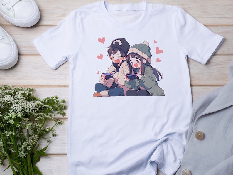May include: A white t-shirt with a graphic print of two anime characters, a boy and a girl, playing video games. The boy is wearing a blue and white hat and a brown sweater. The girl is wearing a green sweater and a white hat. The graphic is surrounded by pink hearts.
