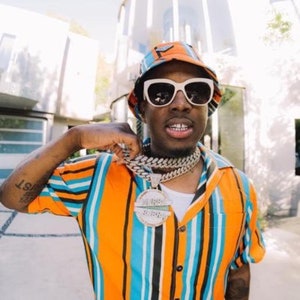 May include: A person wearing a white bucket hat with a black and orange striped pattern, white sunglasses, and a gold chain with a large pendant. The person is wearing a striped orange and blue button-down shirt.