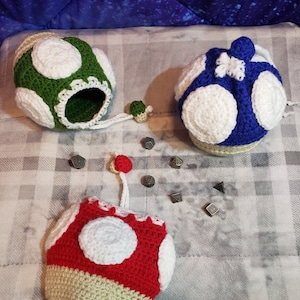 May include: Three crocheted dice bags shaped like Super Mario mushrooms. The bags are green, blue, and red with white spots. There are several silver dice scattered around the bags.