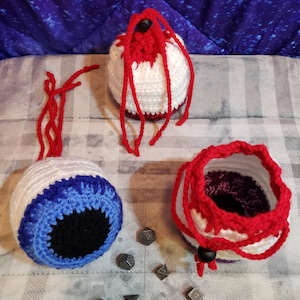 May include: Three crocheted dice bags, one with a white and red eye design, one with a white and blue eye design, and one with a white, red, and purple design. The bags are on a white and gray patterned surface with metal dice scattered around them.