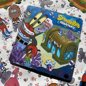 Può includere: Un raccoglitore colorato con personaggi e immagini della serie animata Spongebob Squarepants. La copertina mostra Mr. Krabs, il ristorante Krusty Krab ed elementi sottomarini. Il raccoglitore è circondato da adesivi di cartoni animati.