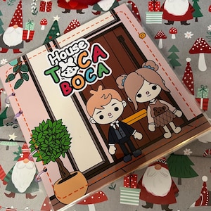 Pode incluir: Um caderno espiral com o texto "House of Toca Boca" e personagens de desenhos animados. A capa tem uma borda rosa e um desenho de uma porta. O fundo é um padrão cinza com tema de Natal com gnomos, árvores e presentes.