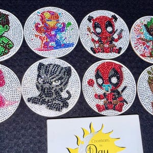 May include: Set of eight round coasters featuring various superhero characters, each embellished with sparkling rhinestones. Characters include Hulk, Iron Man, Deadpool, and Groot, among others. The coasters are white with a rhinestone border.