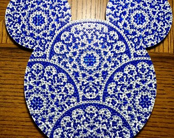 May include: A set of three blue and white decorative coasters in the shape of Mickey Mouse's head. The coasters feature a detailed, ornate pattern with a floral design. The design is made of small, sparkling elements, creating a textured appearance.
