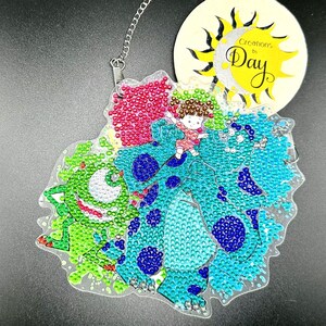 May include: A clear, diamond-painted ornament featuring a cartoon character riding a blue and green creature. The ornament has a chain for hanging and a circular tag with the text "Creations by Day" and a sun design.