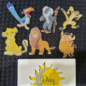 May include: Six diamond art stickers featuring animated animal characters including a bird, a monkey, a meerkat, a lion cub, an adult lion, and a warthog, positioned above a white rectangular sign with a yellow sun logo and the text Creations by Day.