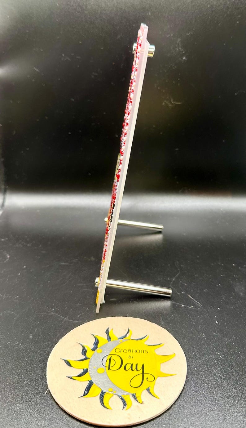 May include: A white picture frame with a red and white design. The frame is standing on a small metal stand. The frame is decorated with rhinestones. The frame is sitting on a brown coaster with a yellow sun and moon design and the text "Creations by Day".