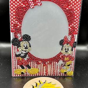 May include: A red and white picture frame with a Mickey Mouse and Minnie Mouse design. The frame is decorated with rhinestones and has a clear oval opening for a photo. The frame is made by Creations by Day.