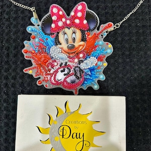 May include: A Minnie Mouse pendant necklace with a silver chain. The pendant features a colorful design of Minnie Mouse with a red bow and a vibrant background. A small canvas with the text "Creations by Day" is also in the image.