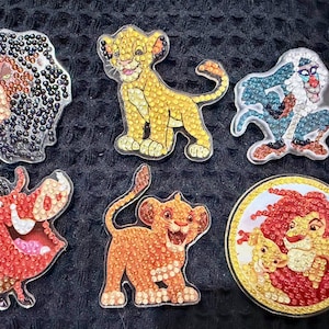 May include: Six diamond art stickers featuring characters from The Lion King, including Simba, Rafiki, and Pumbaa. The stickers are arranged next to a ruler showing a scale in inches, with the largest circular piece measuring about 2.5 inches wide.