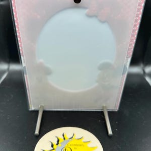 May include: A clear acrylic picture frame with a white background and a pink polka dot border. The frame has a hole at the top and two silver metal stands. The frame is sitting on a coaster with a yellow sun and a silver moon with the text "Greetings of the Day".