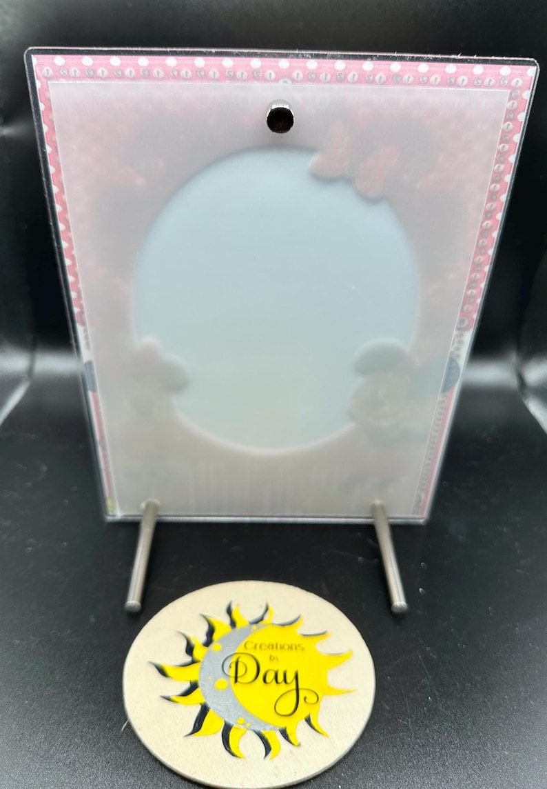 May include: A clear acrylic picture frame with a white background and a pink polka dot border. The frame has a hole at the top and two silver metal stands. The frame is sitting on a coaster with a yellow sun and a silver moon with the text "Greetings of the Day".