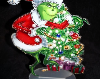 May include: A decorative acrylic standee featuring a green character wearing a red and white Santa suit and hat, holding a decorated green Christmas tree with colorful ornaments and gifts at the base, all embellished with sparkling rhinestones.